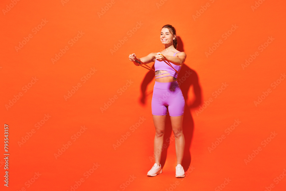 Full body strong young fitness trainer instructor sporty woman sportsman wear purple top clothes in home gym hold elastic rubber band isolated on plain orange background Workout sport fit abs concept Full body strong young fitness trainer instructor sporty woman sportsman wear purple top clothes in home gym hold elastic rubber band isolated on plain orange background Workout sport fit abs concept