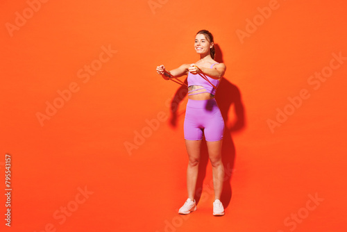 Full body strong young fitness trainer instructor sporty woman sportsman wear purple top clothes in home gym hold elastic rubber band isolated on plain orange background Workout sport fit abs concept