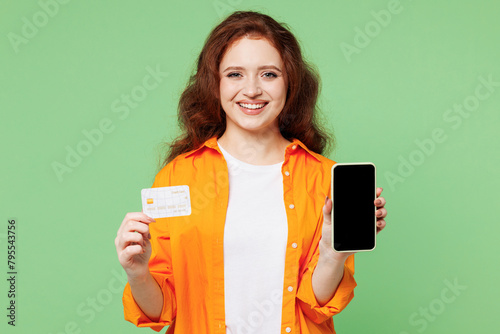 Young woman wears orange shirt white t-shirt casual clothes using blank screen mobile cell phone hold credit bank card shopping online order delivery booking tour isolated on plain green background.