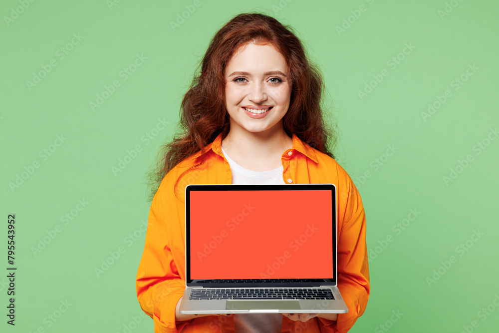 Naklejka premium Young smiling happy ginger IT woman wear orange shirt white t-shirt casual clothes hold use work on blank screen laptop pc computer isolated on plain pastel light green background. Lifestyle concept.
