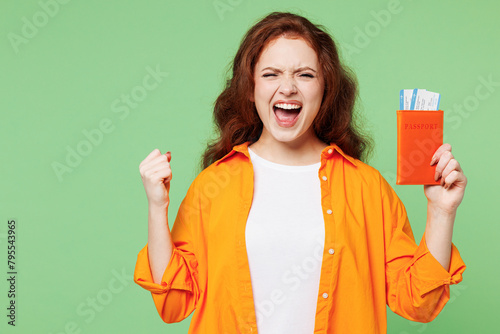 Traveler woman wear orange casual clothes hold passport ticket do winner gesture isolated on plain green background. Tourist travel abroad in free spare time getaway. Air flight trip journey concept. #795543965