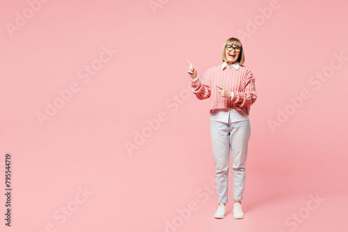 Full body elderly happy woman 50s years old wear sweater shirt casual clothes glasses point index finger aside on area isolated on plain pastel light pink background studio portrait Lifestyle concept