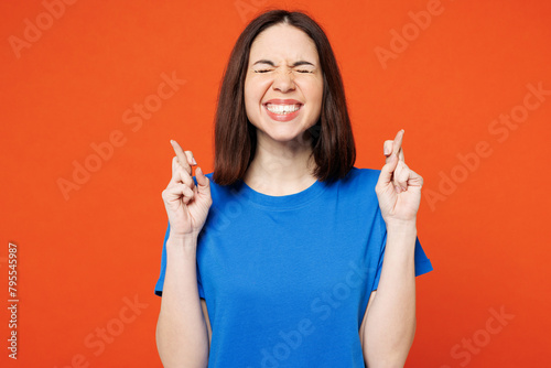 Young smiling happy woman wear blue t-shirt casual clothes waiting for special moment, keeping fingers crossed, making wish isolated on plain red orange background studio portrait. Lifestyle concept. photo