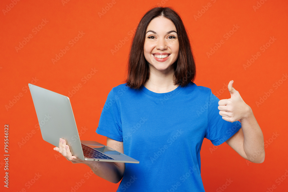 Young smiling satisfied happy IT woman she wear blue t-shirt casual clothes hold use work on laptop pc computer show thumb up isolated on plain red orange background studio portrait Lifestyle concept Young smiling satisfied happy IT woman she wear blue t-shirt casual clothes hold use work on laptop pc computer show thumb up isolated on plain red orange background studio portrait Lifestyle concept