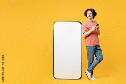 Full body young happy man he wear pink t-shirt casual clothes stand near big huge blank screen mobile cell phone with area use smartphone isolated on plain yellow orange background. Lifestyle concept.