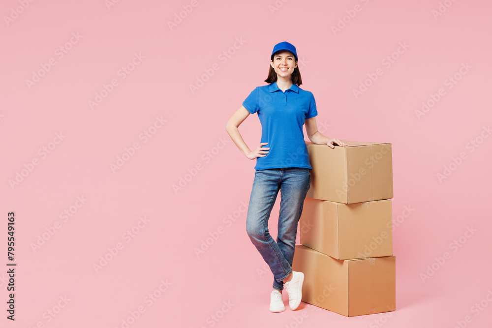 Full body delivery girl employee woman wear blue cap t-shirt uniform workwear work as dealer courier stand near stack of cardboard boxes look camera isolated on plain pink background. Service concept. Full body delivery girl employee woman wear blue cap t-shirt uniform workwear work as dealer courier stand near stack of cardboard boxes look camera isolated on plain pink background. Service concept.