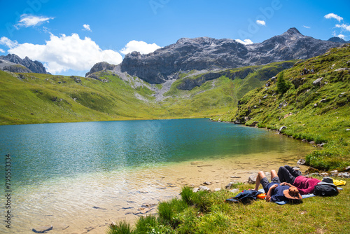 Couple (unrecognizable people) relaxing on the shore of  beautiful La Plagne lake in the French Alps. Other hikers walking up the hill. Peisey valley, Savoie, France. Summer active holidays in nature. photo