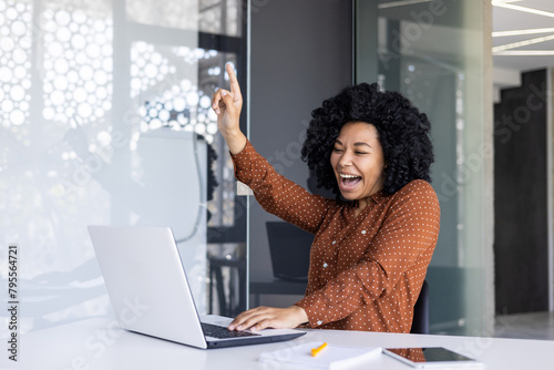 A joyful African American woman in a polka dot shirt celebrating a success or good news at her modern office desk, making a peace sign with enthusiasm.