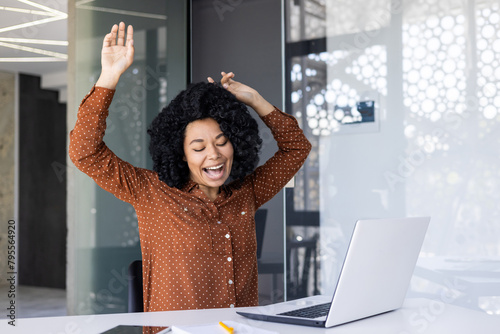 A happy African American woman feeling ecstatic, dancing and singing at her desk in a well-lit, stylish office. She shows pure joy and carefree attitude.