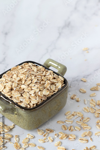 Rolled oat flakes (raw oatmeal) in bowl isolated on white marble table. Close-up. Copy space.