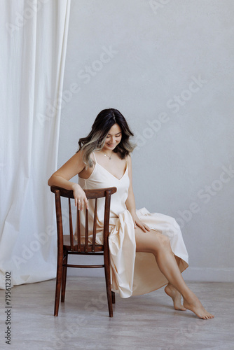 A woman is sitting on a wooden chair in a room with white walls. She is wearing a dress and smiling. Concept of relaxation and comfort, as the woman is at ease in her surroundings