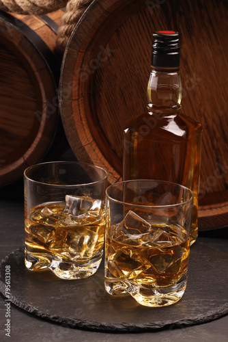 Whiskey with ice cubes in glasses, bottle and wooden barrels on black table