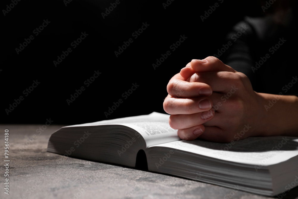 Fototapeta premium Religion. Christian woman praying over Bible at table against black background, closeup. Space for text