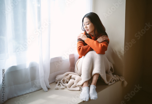 Pensive young Asian woman in warm knitted clothes sitting on windowsill in room. Negative human facial expression.