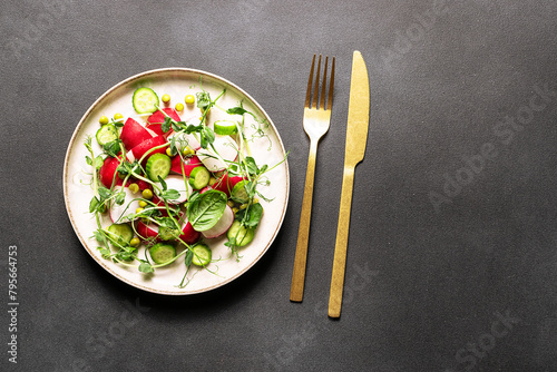 Fresh spring salad with red radish, cucumber,aromatic herbs and olive oil on black stone table close up. Healthy diet food concept.
