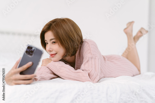 Happy beautiful Asian woman in warm knitted pink clothes taking selfie or recording video vlog on smartphone while lying on bed. Lifestyle with devices addiction and Holiday activity.