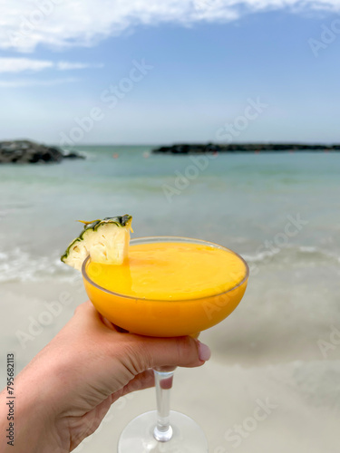 Close-up view of female hand holding friut pineapple cocktail in drinking glass on sandy beach in a spring day. Soft focus. Copy space. Sea resorts and vacations theme.
