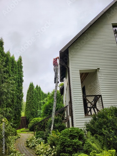 Man on ladder diligently repairing roof of charming white wooden house with determination and hard work under rainy sky, showcasing his craftsmanship in home maintenance.