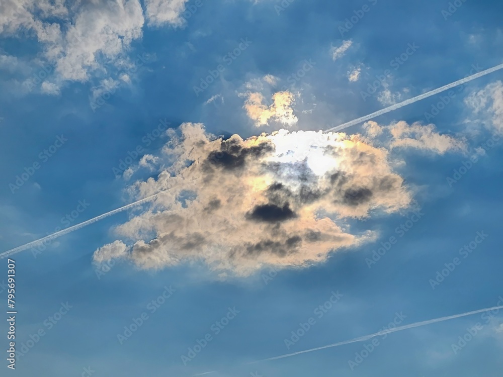 A magnificent cumulus cloud with a radiant silver lining drifts in a vivid blue sky, basking in the warm sun glow, as contrails intersect the heavens, leaving a trail of wonder and awe. A magnificent cumulus cloud with a radiant silver lining drifts in a vivid blue sky, basking in the warm sun glow, as contrails intersect the heavens, leaving a trail of wonder and awe.