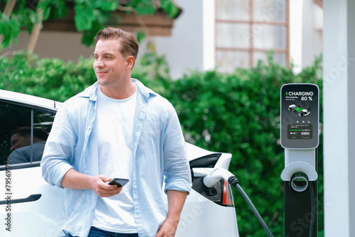 Modern eco-friendly man recharging electric vehicle from home EV charging station. Innovative EV technology utilization for tracking energy usage to optimize battery charging at home. Synchronos photo