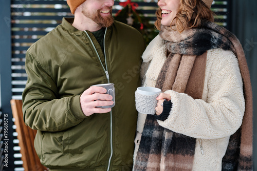Medium crop shot of happy Caucasian couple wearing warm clothes holding mugs standing outdoors on winter day