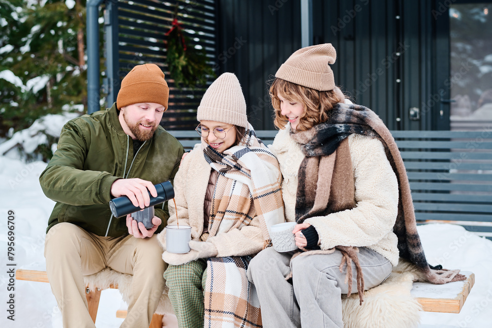 Bearded man pouring hot tea from vacuum flask into mug while sitting on bench outdoors with his wife and daughter