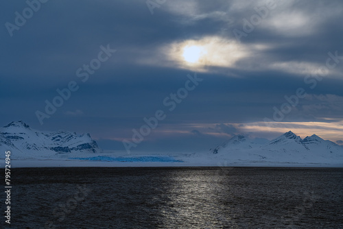 2022-05-08 THE SUN BREAKING THROGUH THE CLOUDS PROVIDING A REFLECTION ON THE WATER WITH SNOW COVERED MOUNTAINS IN SVALBARD NORWAY IN THE ARCTIC