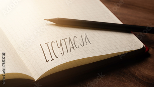  A handwritten inscription "Licytacja" on a grille of an open notebook on a wooden countertop, next to a black pencil, lighting of light. (selective focus), translation: Auction
