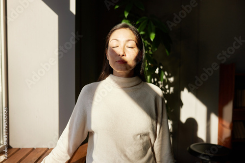 Indoor portrait of beautiful asian girl enjoying morning spring sunbeams kissing her face through window, standing with closed eyes next to windowsill in sweater, feeling relaxed and peaceful