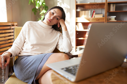 Sad tired pretty asian businesswoman in elegant casual clothes sitting in armchair leaning head on hand and looking at laptop screen with exhausted face, having rest after hard working day