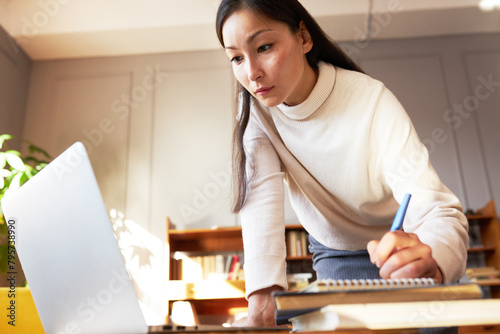 Low view of smart elegant businesswoman standing in front of laptop noting down important information to notepad. Beautiful female secretary looking at screen of computer making notes