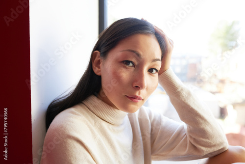 Closeup portrait of gentle charming asian female in sweater sitting on windowsill, having rest admiring city life, leaning head on hand while looking at camera, having rest and relaxing