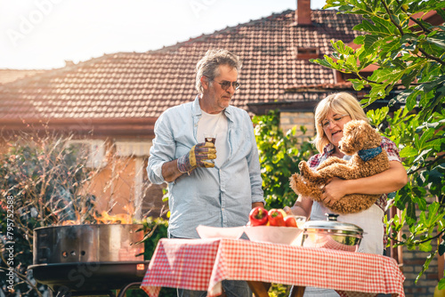 Cheerful senior couple playing with dog while making barbecue in backyard on a sunny day. Woman holding a poodle in the backyard.