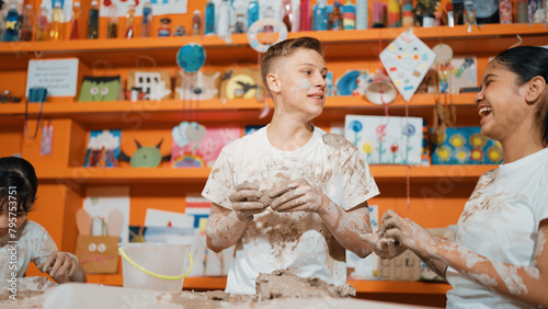 Group of happy diverse children playing clay while wearing muddy shirt at art lesson. Cute highschool girl playing with dough while putting clay on happy boy shirt at pottery workshop. Edification.