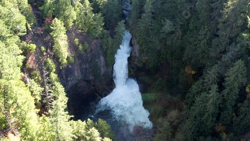 A waterfall is shown in the air with trees in the background. The water is flowing down the waterfall and the trees are green photo