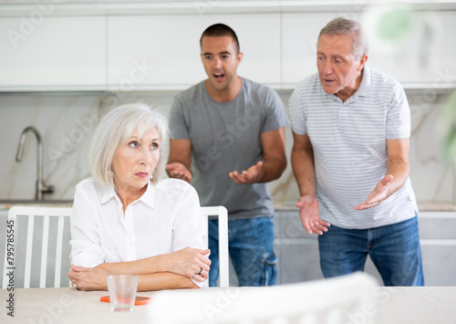 Elderly man and adult man during family quarrel with elderly woman in kitchen