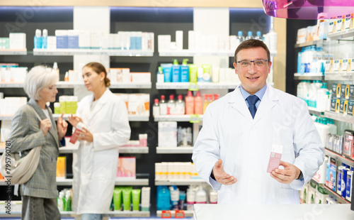 Polite middle-aged male pharmacist demonstrating preparation in box in chemist's shop with large assortment