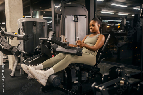 Fototapeta Naklejka Na Ścianę i Meble -  A strong black sportswoman doing workouts at seated leg curl machine at gym.