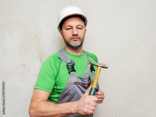 Portrait of a construction worker with tools on light wall background. Builder in grey heavy duty clothes, white safety plastic helmet and green shirt. Male in his 40s with grey beard