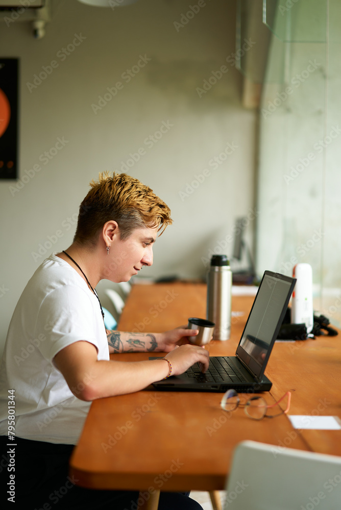 Fotografie Transgender freelancer works on laptop in coworking space, sips tea from vacuum flask mug