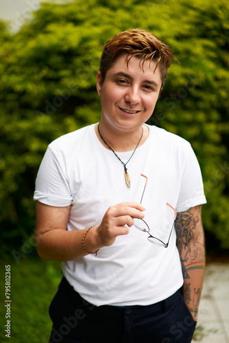 Smiling transgender man outdoors in casual attire, confidence evident in stance with glasses in hand, tattoo visible on arm, plants backdrop suggesting fresh start, inclusivity, gender identity pride.