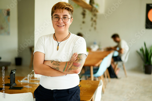 Confident transgender male with tattoos stands in a modern office, smiling colleagues working in the background. He embraces diversity, inclusion in the workplace, wearing casual attire and glasses. photo