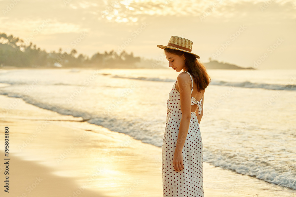 Elegant woman strolls on sunny beach in summer, white polka-dot dress flowing, straw hat on. Relaxed pace, seaside breeze, golden hour light, embodies leisure, vacation spirit near ocean waves.