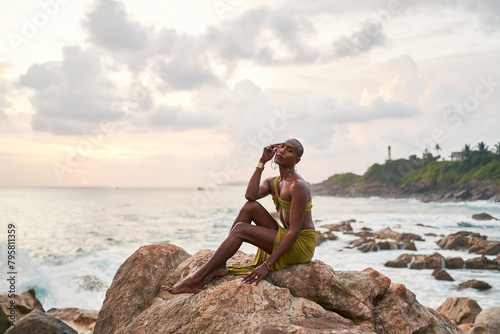 Gay bipoc graceful man poses sitting high on rocks in ocean at sunset. Homosexual slim ethnic fashion model in tropical maxi dress on top of rocky hill above storm at dusk. Pride LGBTQIA