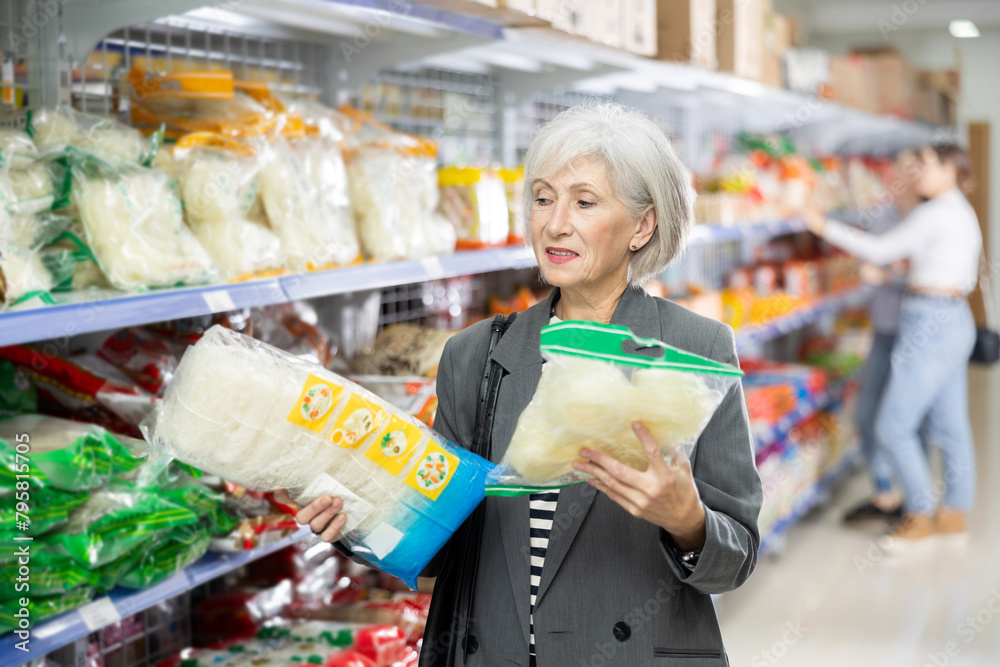 Senior woman in supermarket buys daily groceries, pasta Chinese noodles. Female pensioner buyer examines packaging of product and reads information on label