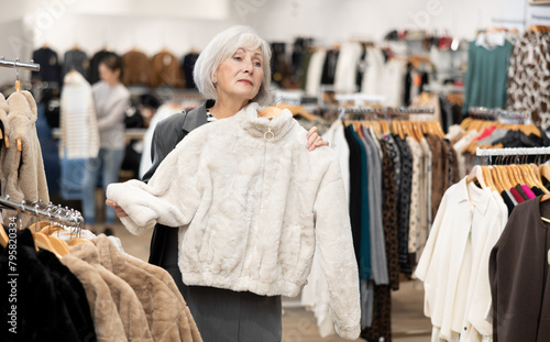 Pleased mature woman choosing short fur coat in clothing shop with large assortment