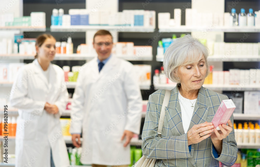 Fototapeta premium Careful mature female customer holding a box with preparation standing against shelves full of medicine in drugstore