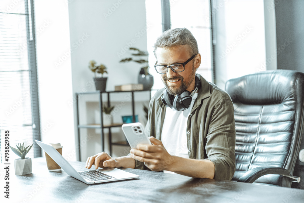A man is sitting at a desk with a laptop and a cell phone. He is smiling and he is enjoying himself A man is sitting at a desk with a laptop and a cell phone. He is smiling and he is enjoying himself