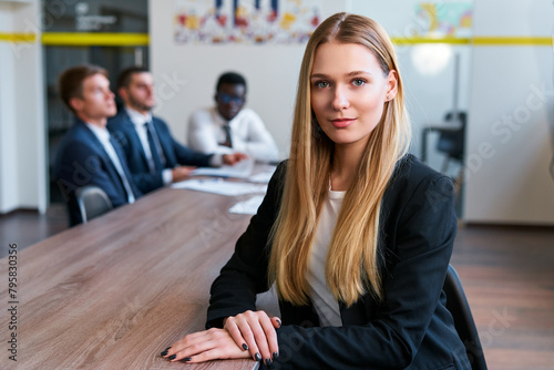 Confident blonde woman sits at boardroom table in modern office, team of male colleagues in background. Professional business team during meeting. Female leader, corporate environment, confident pose. photo