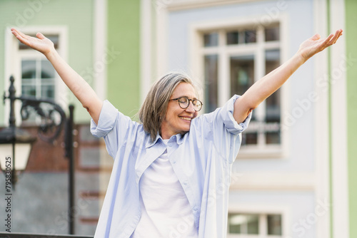 A woman in a blue shirt is standing outside and raising her arms in the air. She is smiling and she is happy
